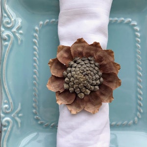 May include: A brown pinecone flower napkin ring on a white napkin, placed on a light blue plate with a decorative border.