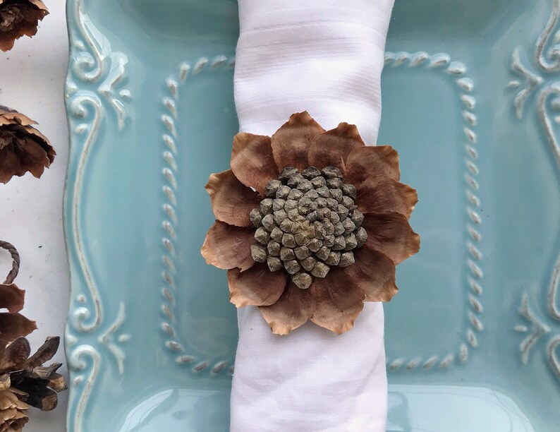 May include: A brown pinecone flower napkin ring on a white napkin, placed on a light blue plate with a decorative border.