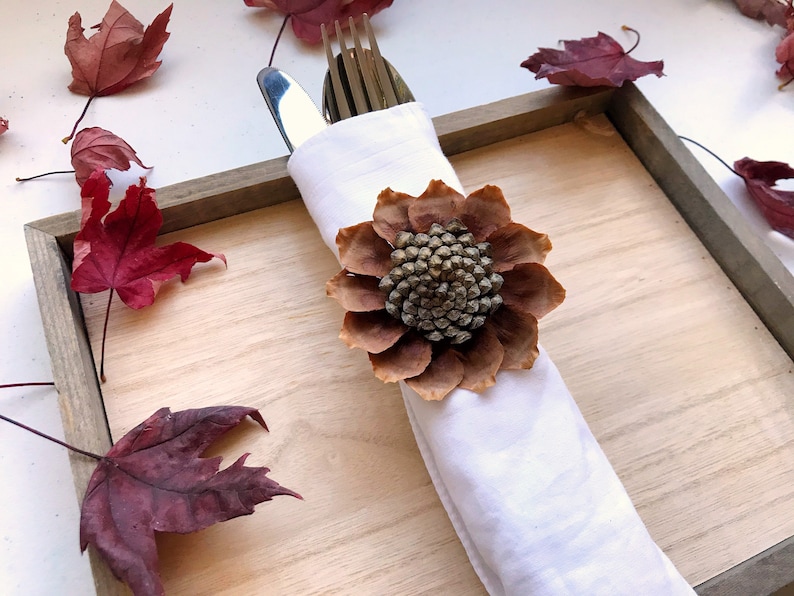 May include: A white napkin with a brown pinecone flower ring, placed on a wooden tray with red fall leaves scattered around it.