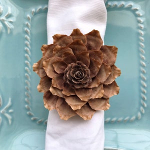 May include: A brown pine cone flower napkin ring. The napkin ring is made from a pine cone and is shaped like a flower. It is sitting on a white napkin and a blue plate.