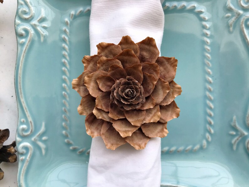 May include: A brown pine cone flower napkin ring. The napkin ring is made from a pine cone and is shaped like a flower. It is sitting on a white napkin and a blue plate.