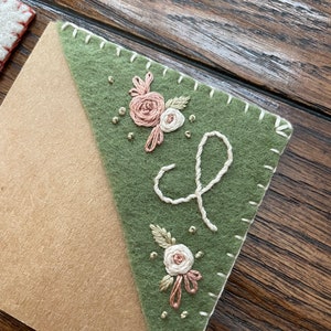 May include: A green felt bookmark with a white stitched border and embroidered flowers and a cursive letter 'L'.