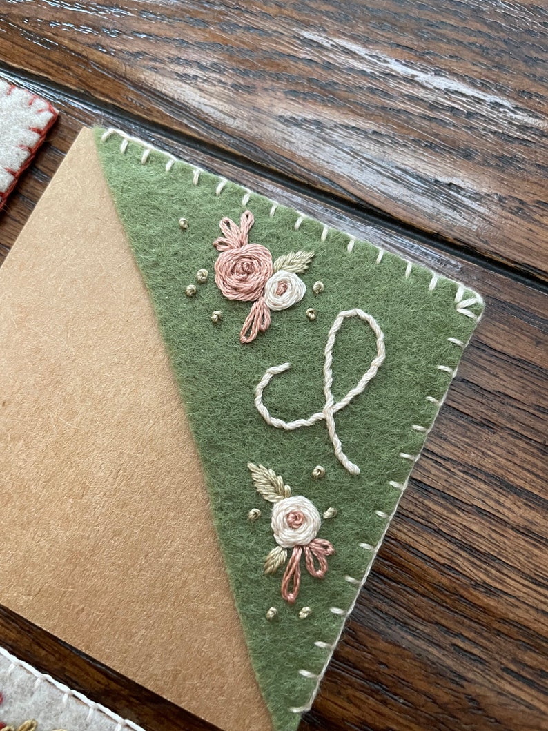 May include: A green felt bookmark with a white stitched border and embroidered flowers and a cursive letter 'L'.