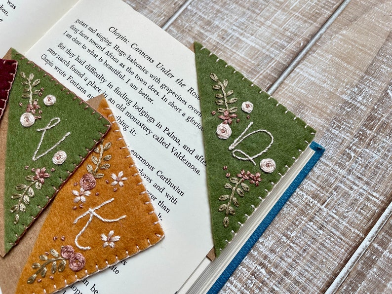 May include: Three felt fabric bookmarks with embroidered floral designs and letters. The bookmarks are in shades of green, brown, and beige. The text on the bookmarks reads "Q", "K", and "V".
