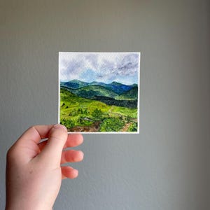 Peut inclure: Petite aquarelle d'un paysage de montagne. L'&oelig;uvre repr&eacute;sente des collines et des montagnes verdoyantes sous un ciel nuageux. La peinture est tenue dans une main sur un fond gris.