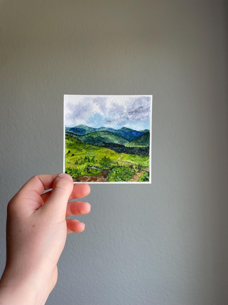 Peut inclure: Petite aquarelle d'un paysage de montagne. L'&oelig;uvre repr&eacute;sente des collines et des montagnes verdoyantes sous un ciel nuageux. La peinture est tenue dans une main sur un fond gris.