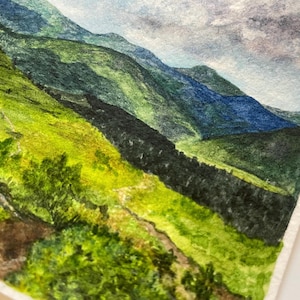 Peut inclure: Aquarelle repr&eacute;sentant un paysage de montagne. L'&oelig;uvre pr&eacute;sente des collines et des montagnes verdoyantes dans des tons de bleu et de vert. Un sentier serpente au premier plan, et le ciel est un m&eacute;lange de bleu et de gris.