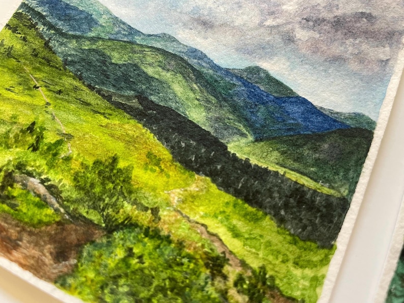 Peut inclure: Aquarelle repr&eacute;sentant un paysage de montagne. L'&oelig;uvre pr&eacute;sente des collines et des montagnes verdoyantes dans des tons de bleu et de vert. Un sentier serpente au premier plan, et le ciel est un m&eacute;lange de bleu et de gris.