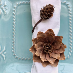 May include: A brown pine cone flower napkin ring on a white napkin, set on a light blue plate with a decorative border.