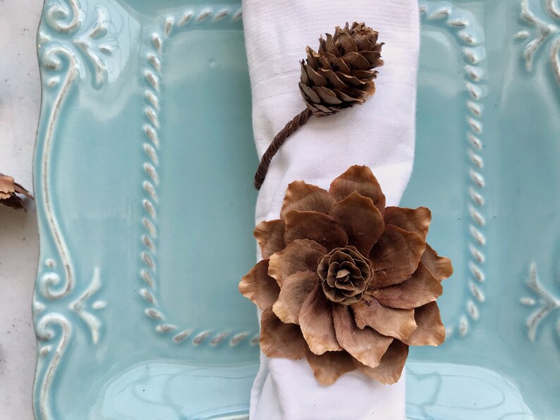 May include: A brown pine cone flower napkin ring on a white napkin, set on a light blue plate with a decorative border.