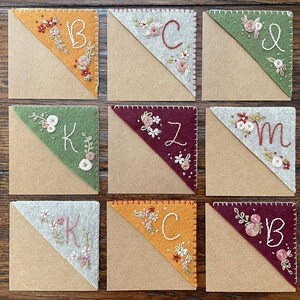 May include: Nine felt corner bookmarks with embroidered floral designs and initials. The bookmarks are in various colors, including orange, green, red, and gray. The initials are K, B, C, Z, and M.