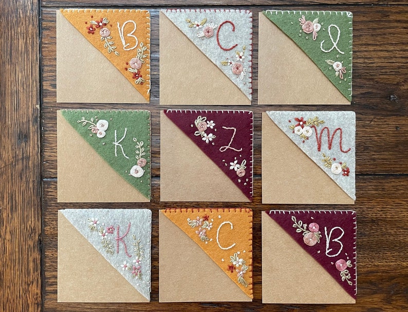 May include: Nine felt corner bookmarks with embroidered floral designs and initials. The bookmarks are in various colors, including orange, green, red, and gray. The initials are K, B, C, Z, and M.