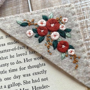 May include: A handmade felt bookmark with embroidered flowers. The corner bookmark features a floral design with red and white roses, green leaves, and small gold accents. The bookmark is placed on a book with printed text.
