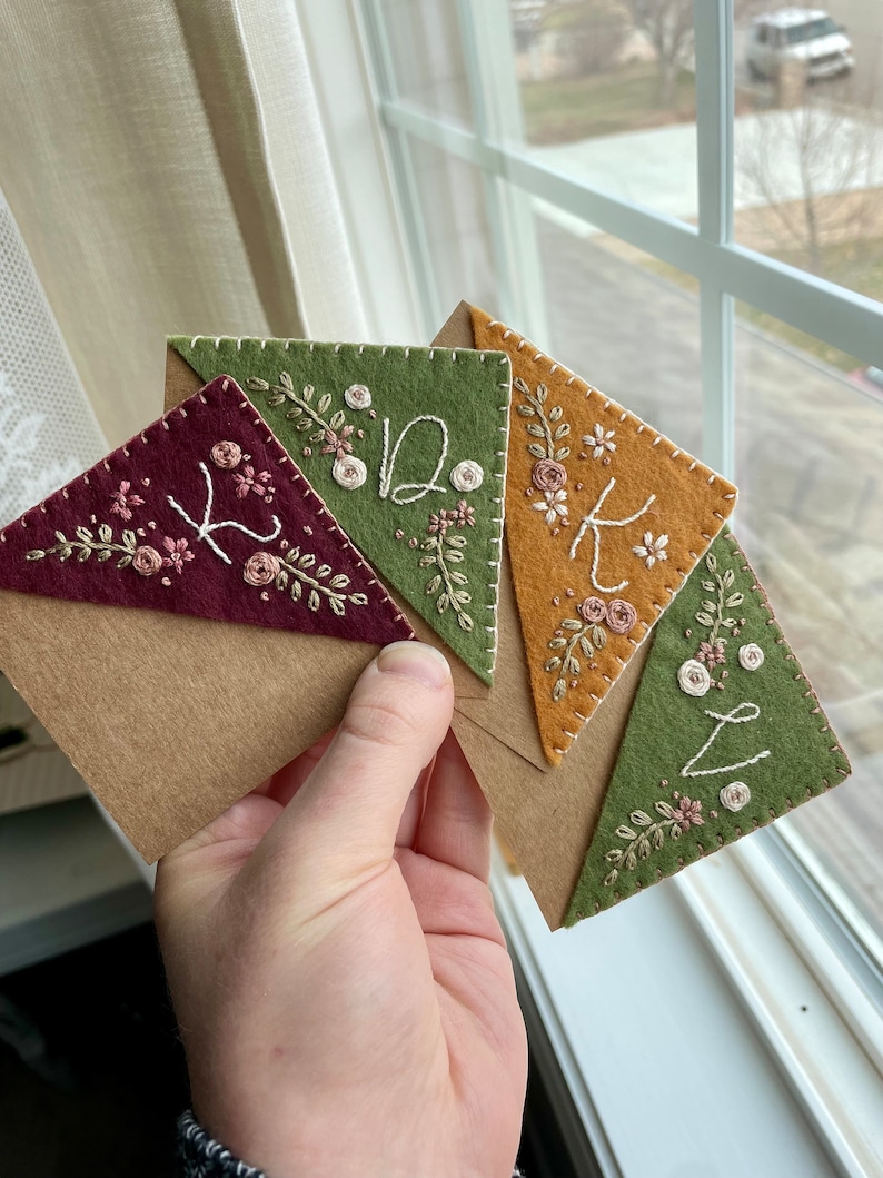 May include: Four felt triangle bookmarks with embroidered floral designs and letters K, D, K, and L. The bookmarks are arranged in a stack and are held in a hand.