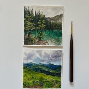 Peut inclure: Deux aquarelles de paysages. L'une repr&eacute;sente un lac turquoise avec des arbres &agrave; feuilles persistantes et des montagnes. L'autre montre des collines verdoyantes sous un ciel nuageux. Un pinceau marron avec une pointe dor&eacute;e est &agrave; c&ocirc;t&eacute; des peintures.