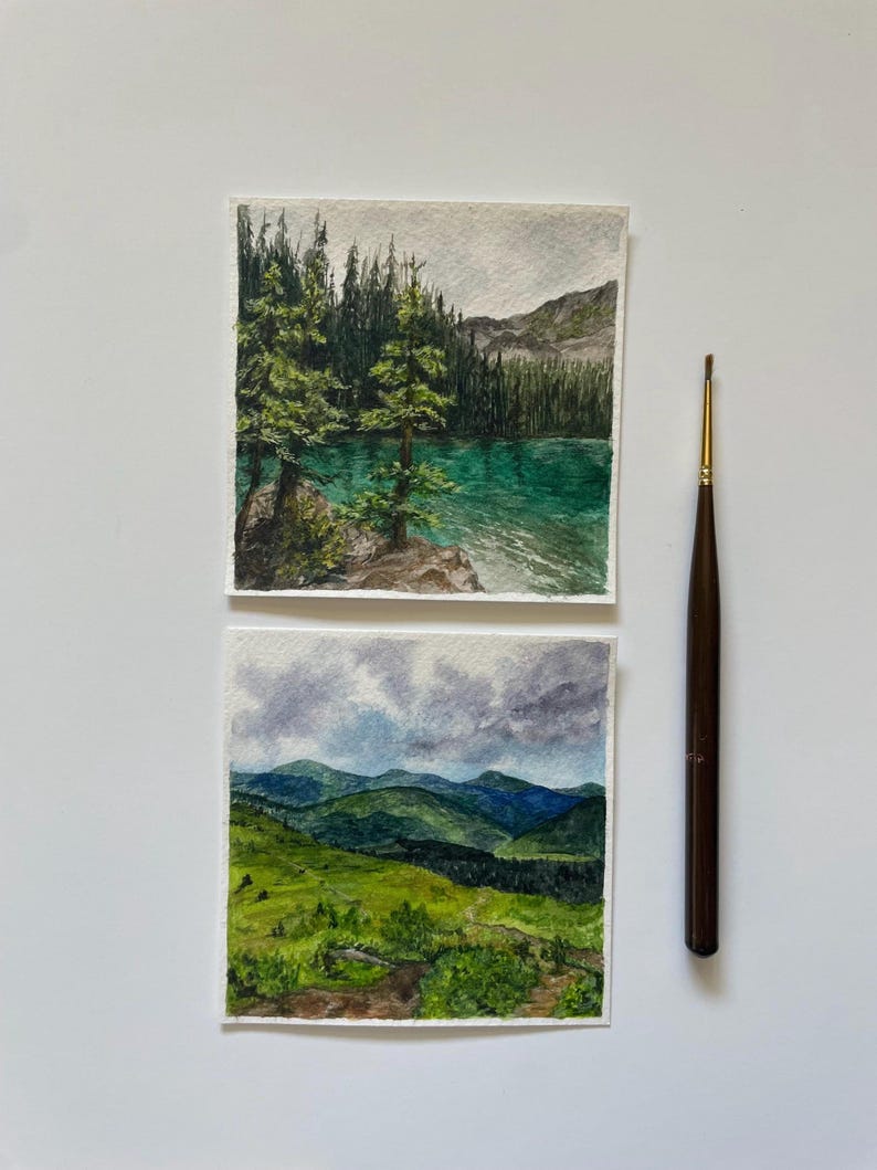 Peut inclure: Deux aquarelles de paysages. L'une repr&eacute;sente un lac turquoise avec des arbres &agrave; feuilles persistantes et des montagnes. L'autre montre des collines verdoyantes sous un ciel nuageux. Un pinceau marron avec une pointe dor&eacute;e est &agrave; c&ocirc;t&eacute; des peintures.