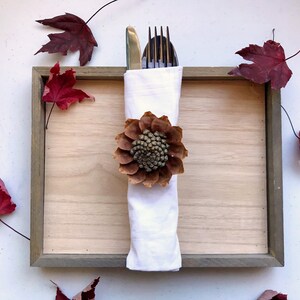 May include: A white napkin with a brown pinecone flower ring, a gold knife and silver fork are placed inside the napkin. The napkin is on a wooden tray with a gray frame. There are red maple leaves scattered around the tray.