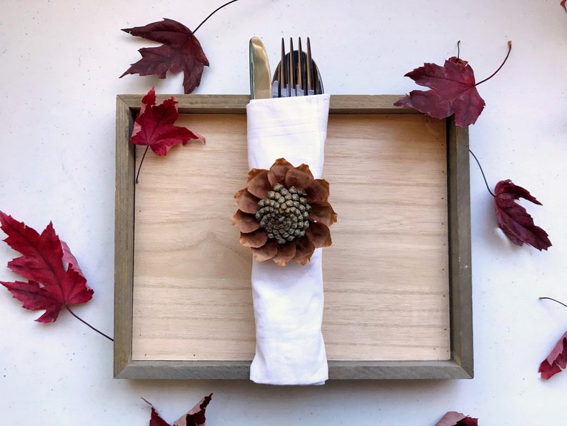 May include: A white napkin with a brown pinecone flower ring, a gold knife and silver fork are placed inside the napkin. The napkin is on a wooden tray with a gray frame. There are red maple leaves scattered around the tray.