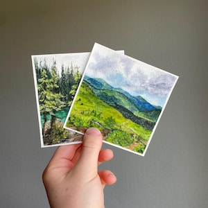 Peut inclure: Deux petites peintures &agrave; l'aquarelle de paysages. L'une repr&eacute;sente une for&ecirc;t avec un lac turquoise, l'autre une cha&icirc;ne de montagnes avec des collines vertes et un ciel nuageux. Les peintures sont tenues sur un fond gris.