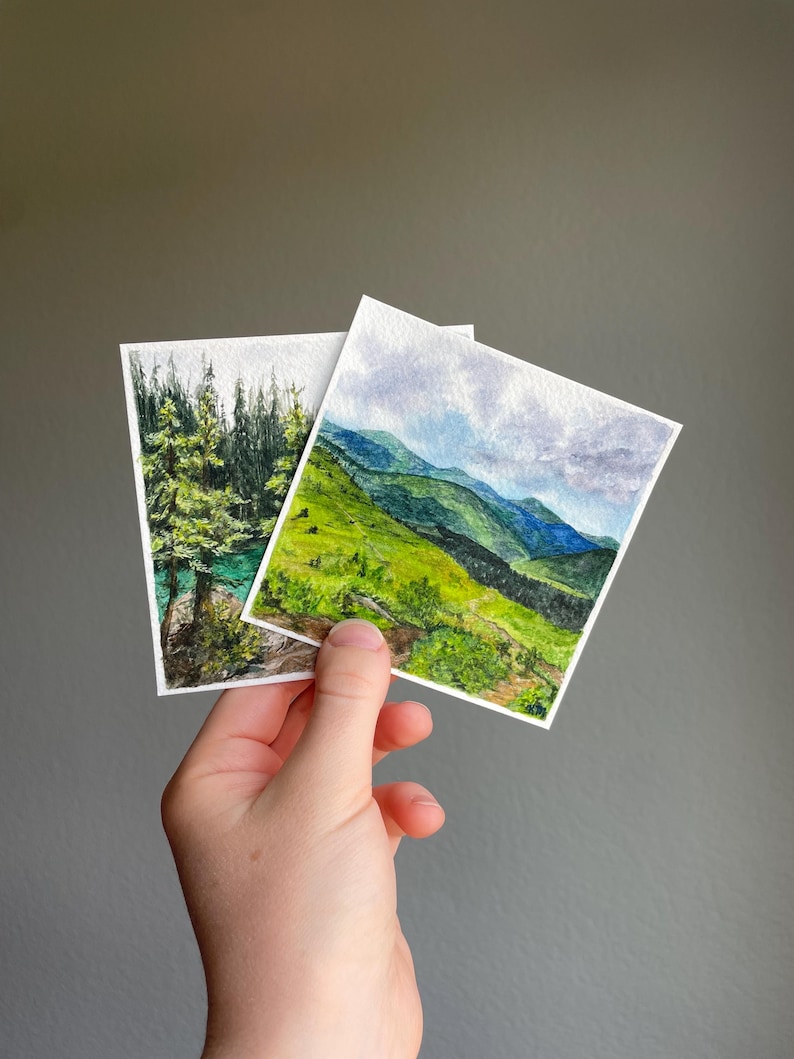 Peut inclure: Deux petites peintures &agrave; l'aquarelle de paysages. L'une repr&eacute;sente une for&ecirc;t avec un lac turquoise, l'autre une cha&icirc;ne de montagnes avec des collines vertes et un ciel nuageux. Les peintures sont tenues sur un fond gris.