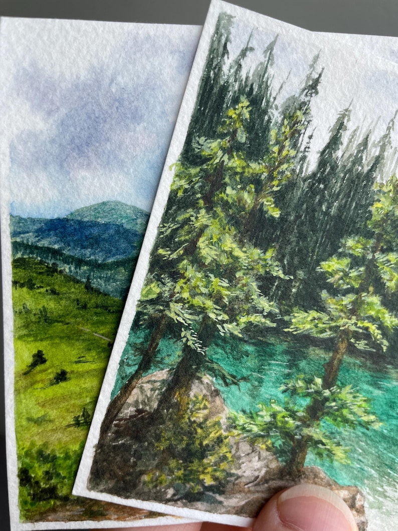 Peut inclure: Deux peintures &agrave; l'aquarelle de paysages. L'une repr&eacute;sente un champ vert et des montagnes bleues sous un ciel nuageux. L'autre montre des arbres verts et de l'eau turquoise. Les deux sont sur papier blanc.