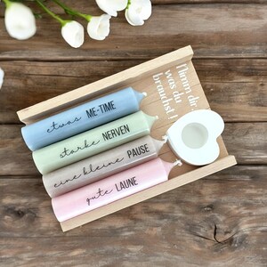 Könnte beinhalten: Ein Holztablett mit fünf Kerzen, jede in einer anderen Farbe und mit einem Spruch darauf. Die Sprüche lauten "etwas ME-TIME", "starke NERVEN", "eine kleine PAUSE", "gute LAUNE" und "Nimm dir, was du brauchst!" Das Tablett hat außerdem einen kleinen weißen herzförmigen Kerzenhalter.