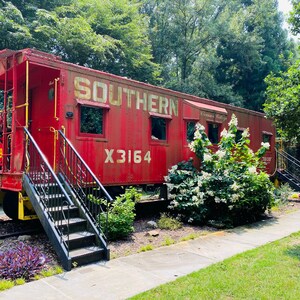 May include: A vintage red Southern caboose with gold lettering and the number X3164. The caboose has black stairs and is surrounded by greenery, including trees and bushes. The image is taken outdoors.
