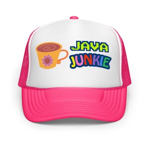 以下が含まれることがあります： 白いフロントパネルにコーヒーカップのグラフィックとカラフルなピクセル化されたテキストで「JAVA JUNKIE」という文字が入った、ホットピンクのトラッカーハット。帽子はメッシュバックと湾曲したつばが付いています。
