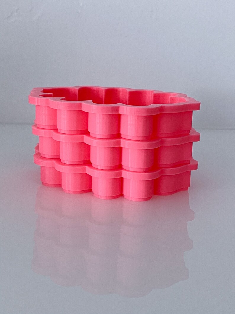 May include: A stack of three bright pink, cloud-shaped cookie cutters. Each cutter has a scalloped edge and a cylindrical design. The cutters are made of plastic and sit on a reflective surface.