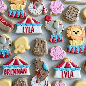 May include: A collection of colorful circus-themed sugar cookies decorated with various designs, including a lion, elephant, bear, hot dog, and a tent with the names "Brennan" and "Lyla".