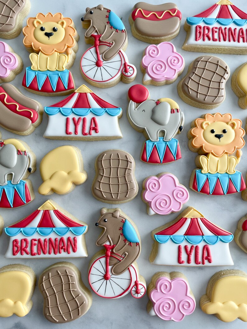 May include: A collection of colorful circus-themed sugar cookies decorated with various designs, including a lion, elephant, bear, hot dog, and a tent with the names "Brennan" and "Lyla".