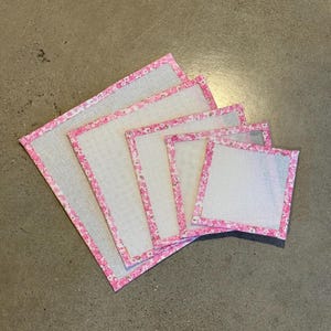 Puede incluir: Cinco cuadrados de lona blanca de diferentes tamaños con ribete floral rosa. Los cuadrados están dispuestos en orden de tamaño graduado, con el cuadrado más grande en la parte inferior y el cuadrado más pequeño en la parte superior.