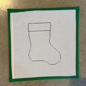 May include: A square needlepoint canvas with a green border. The canvas has a white background and a grey outline of a Christmas stocking. The canvas is designed for needlepoint projects.