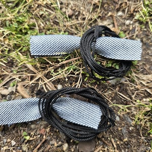 May include: Two black cords with white and blue striped fabric wrapped around them. The cords are lying on a bed of brown dirt and green grass.