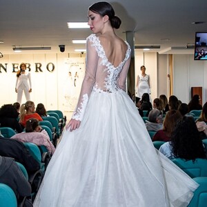 Puede incluir: Un vestido de novia blanco con falda de tul y mangas largas. El vestido presenta delicados detalles de encaje en las mangas y la espalda. La modelo est&aacute; en una pasarela durante un desfile de moda.
