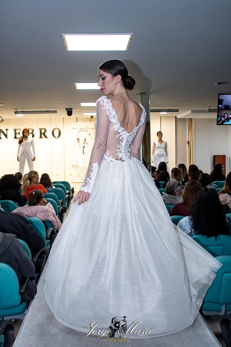 Puede incluir: Un vestido de novia blanco con falda de tul y mangas largas. El vestido presenta delicados detalles de encaje en las mangas y la espalda. La modelo est&aacute; en una pasarela durante un desfile de moda.