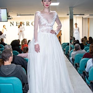 Puede incluir: Una modelo con un vestido de novia blanco con detalles de encaje floral en el corpi&ntilde;o y mangas largas desfila por una pasarela. El vestido tiene una falda amplia y fluida. El evento es un desfile de moda, con p&uacute;blico sentado en sillas azules.