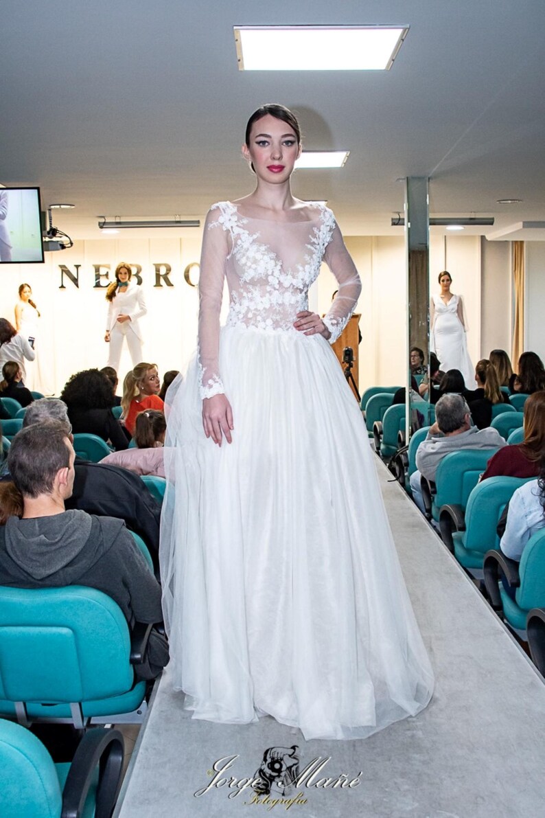 Puede incluir: Una modelo con un vestido de novia blanco con detalles de encaje floral en el corpi&ntilde;o y mangas largas desfila por una pasarela. El vestido tiene una falda amplia y fluida. El evento es un desfile de moda, con p&uacute;blico sentado en sillas azules.
