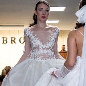 Puede incluir: Vestido de novia blanco con corpi&ntilde;o transparente y detalles de encaje floral. El vestido tiene mangas largas y una falda amplia. El vestido es elegante y adecuado para una boda. La modelo est&aacute; frente a un fondo borroso.