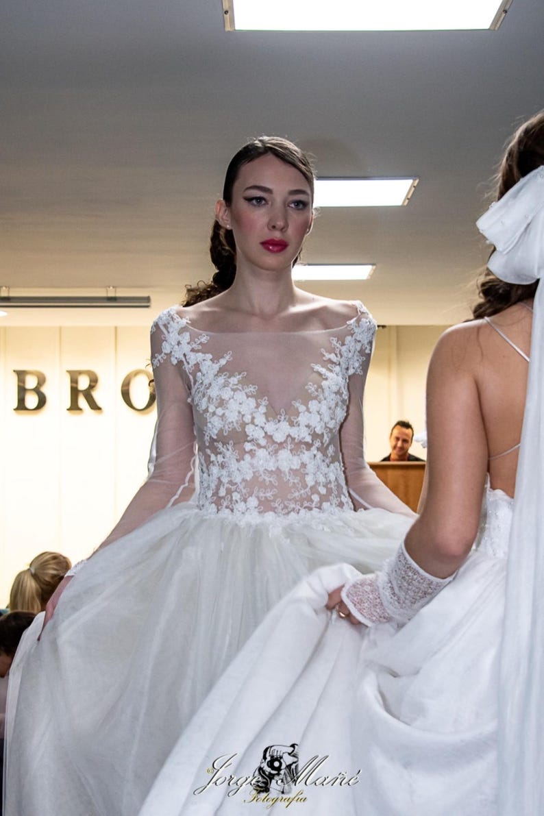 Puede incluir: Vestido de novia blanco con corpi&ntilde;o transparente y detalles de encaje floral. El vestido tiene mangas largas y una falda amplia. El vestido es elegante y adecuado para una boda. La modelo est&aacute; frente a un fondo borroso.