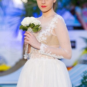 Puede incluir: Vestido de novia blanco con falda de tul y detalles de encaje en el corpi&ntilde;o y las mangas. La novia sostiene un ramo de rosas blancas. El vestido tiene un escote transparente y mangas largas.