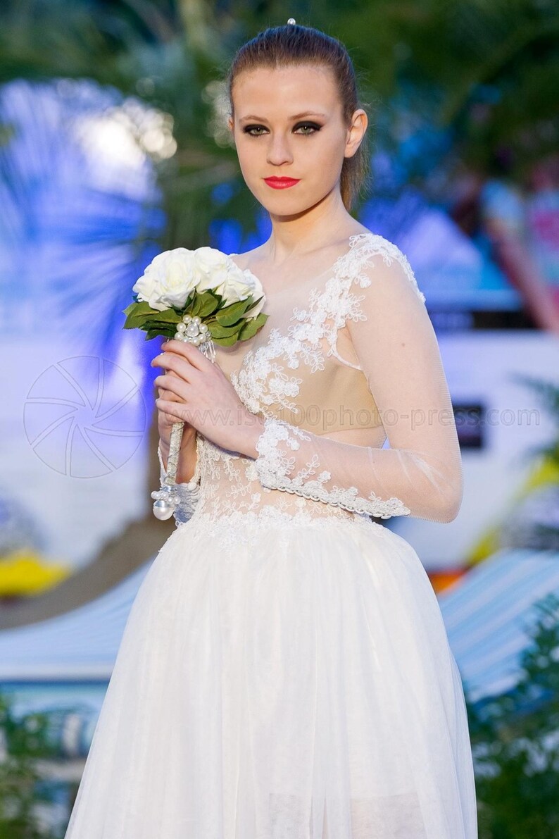 Puede incluir: Vestido de novia blanco con falda de tul y detalles de encaje en el corpi&ntilde;o y las mangas. La novia sostiene un ramo de rosas blancas. El vestido tiene un escote transparente y mangas largas.