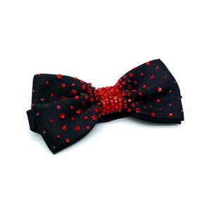 May include: A black bow tie with red rhinestone embellishments. The bow tie has a center cluster of rhinestones and smaller rhinestones scattered across the fabric.