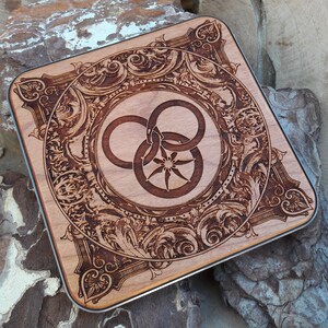 Puede incluir: Un posavasos de madera con un dise&ntilde;o intrincado y ornamentado que presenta un s&iacute;mbolo estilizado de tres anillos entrelazados. El s&iacute;mbolo est&aacute; rodeado por un borde decorativo de hojas y enredaderas en espiral.