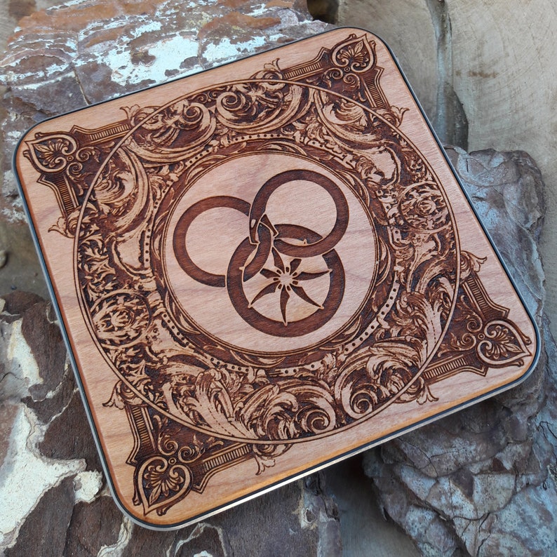 Puede incluir: Un posavasos de madera con un dise&ntilde;o intrincado y ornamentado que presenta un s&iacute;mbolo estilizado de tres anillos entrelazados. El s&iacute;mbolo est&aacute; rodeado por un borde decorativo de hojas y enredaderas en espiral.