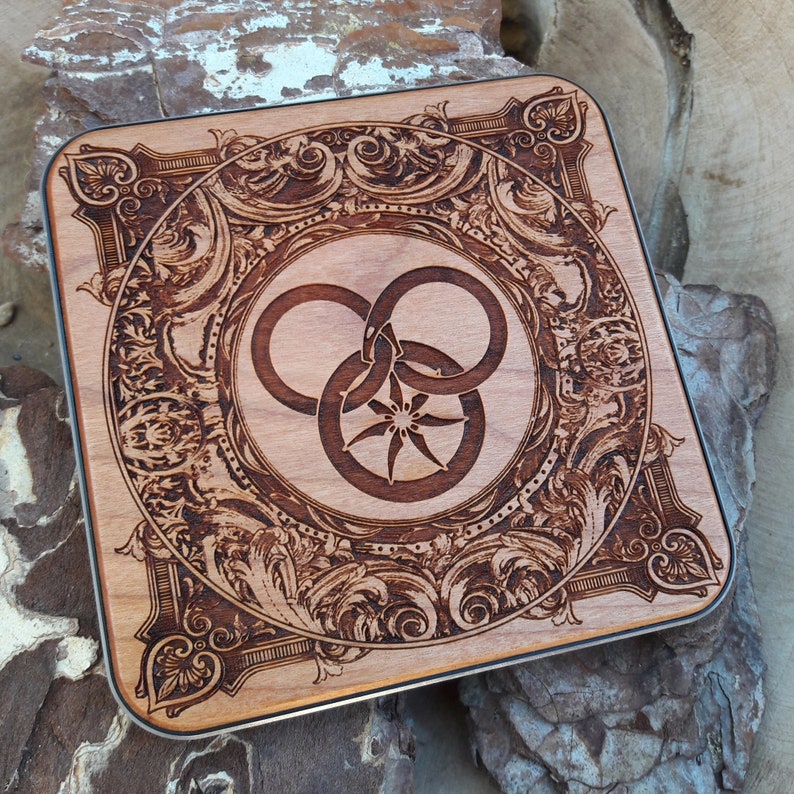 Puede incluir: Un posavasos de madera con un dise&ntilde;o intrincado y ornamentado que presenta un s&iacute;mbolo estilizado de tres c&iacute;rculos entrelazados con una estrella en el centro. El posavasos est&aacute; hecho de madera oscura con un acabado natural.