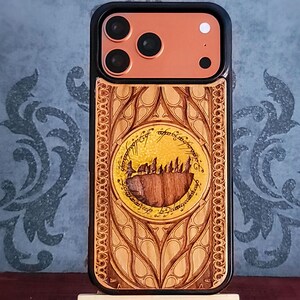May include: A black smartphone case featuring a copper-colored camera module and a light wood back panel. The wood is laser-engraved with an intricate circular design containing a mountain silhouette and Elvish script, surrounded by ornate patterns.