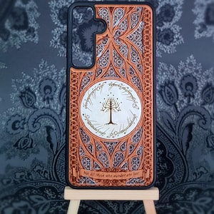 Peut inclure: Coque de téléphone ornée d'un motif détaillé dans des tons bruns et beiges. Elle présente un emblème circulaire central avec un arbre et une écriture, ainsi que le texte "Not all those who wander are lost". Bordure noire.