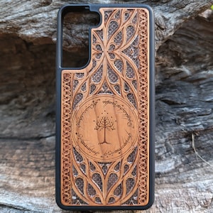 Może przedstawiać: Drewniane etui na telefon z misternymi rzeźbionymi wzorami. Etui posiada centralny okrągły emblemat z drzewem i napisem, otoczony szczegółowymi wzorami. Etui ma czarną obwódkę i jest wykonane z jasnobrązowego drewna.