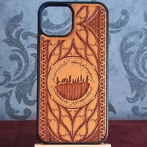 May include: A light brown wooden phone case with a black border and a large camera cutout. The back features an intricate laser-engraved design of a group of figures walking on a ridge, surrounded by a circular border with elvish-style script.